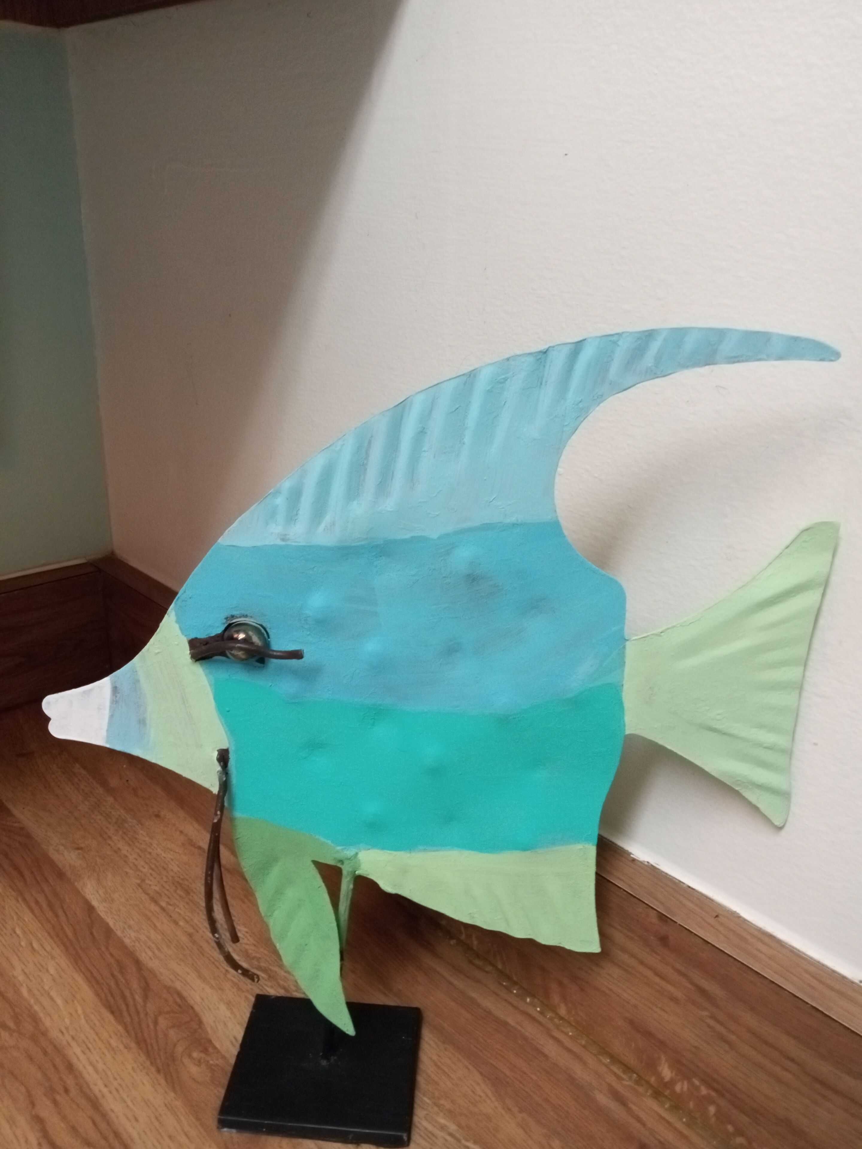 Une sculpture en forme de poisson, posée sur une base noire, elle-même placée sur un plancher en bois.