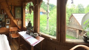 A wooden cabin with a table and chairs, a window with a view of greenery and a wooden structure outside.