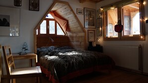 A cozy bedroom with a sloped ceiling, a bed with a patterned bedspread, a wooden chair, a small Christmas tree, and a window with string lights.
