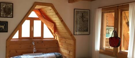 Een gezellige slaapkamer met een houten schuine plafond, een bed met een bloemenprint bedspread, een raam met gordijnen, en een radiator.