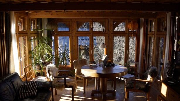A cozy wooden interior with a round table, chairs, and a sofa. Large windows allow natural light and views of the outside.