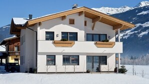 A modern two-story house with a wooden balcony, large windows, and a snowy mountain backdrop.