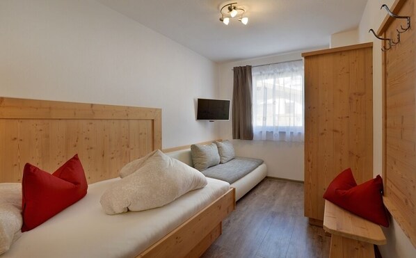 A room with a wooden bed, a sofa, a TV mounted on the wall, and a window with curtains.