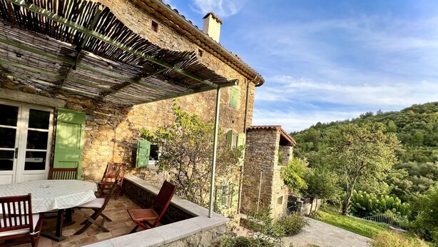 A stone building with a patio area featuring a table and chairs, surrounded by greenery and a view of a hillside.