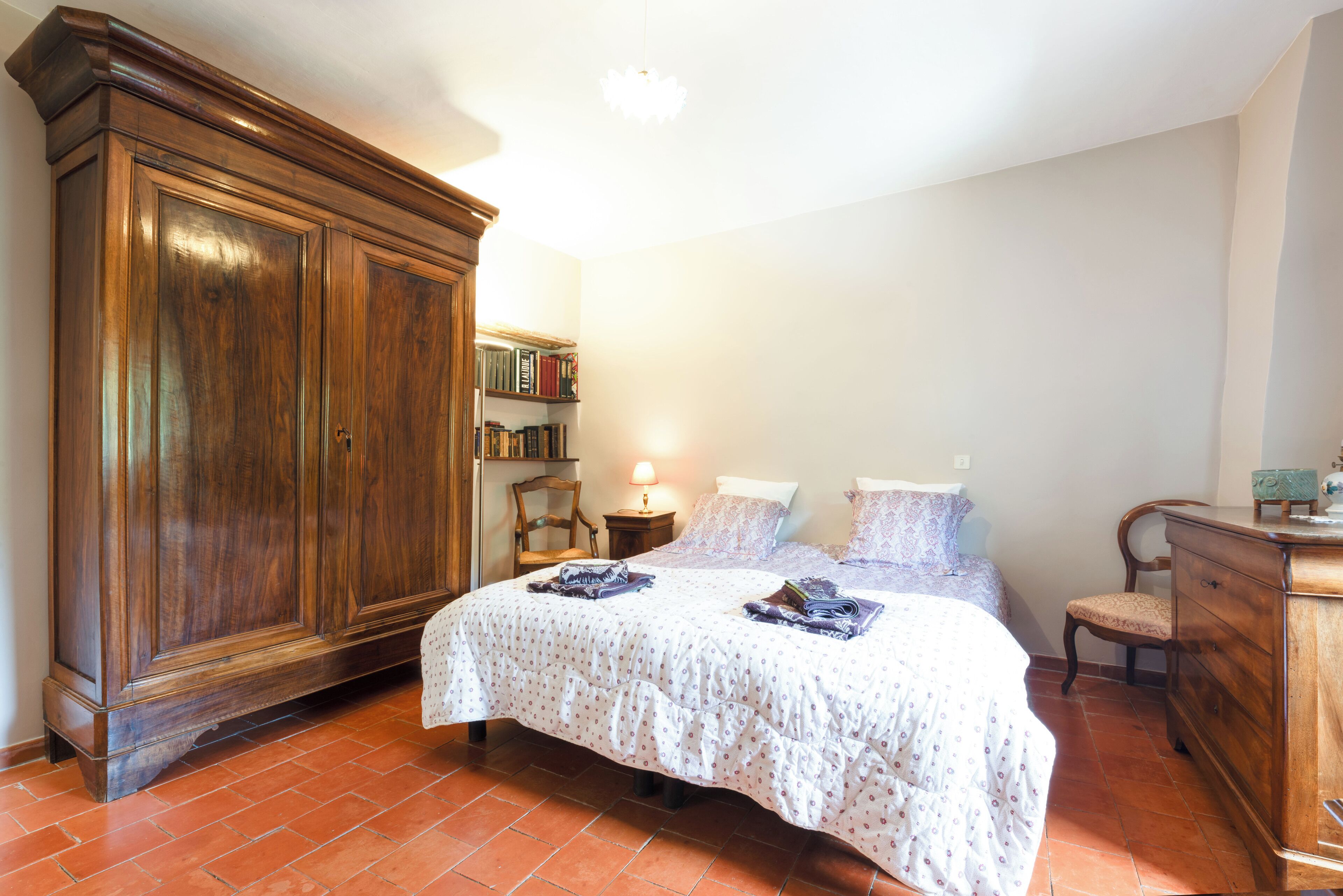 A bedroom with a large wooden wardrobe, a bed with a patterned bedspread, a nightstand with a lamp, and a chair.
