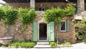 A stone building with a rustic entrance, green shutters, and a potted plant.