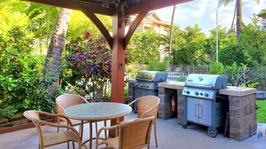 An outdoor patio with wicker chairs, a glass table, and two barbecue grills.