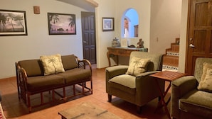 A cozy living room with a brown sofa, two armchairs, and a wooden coffee table. There are framed pictures on the wall, a small table with decorative items, and a door leading to another room. - Villa Sunset, Ocean View rooftop, just 3 blocks from the beach (San Pancho)