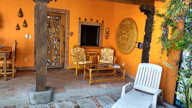 A cozy living area with a wooden door, two patterned chairs, a white plastic chair, a small table, and a decorative wall piece. - Casa de Como Casita #2 - Ajijic Village (Ajijic)