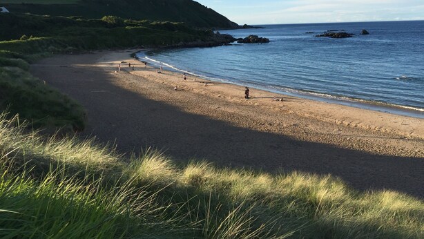 Een kuststrand met een golvende oever, begroeide duinen en rotsachtige uitstulpingen.