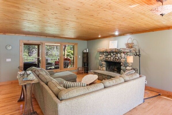 A cozy living room with a stone fireplace, wooden ceiling, and large windows overlooking a forest.