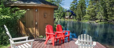 A wooden deck with two rocking chairs and a table, overlooking a calm lake surrounded by trees.
