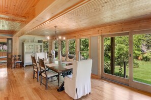 A spacious dining area with a wooden table and chairs, large windows offering a view of the outdoors, and a modern chandelier.