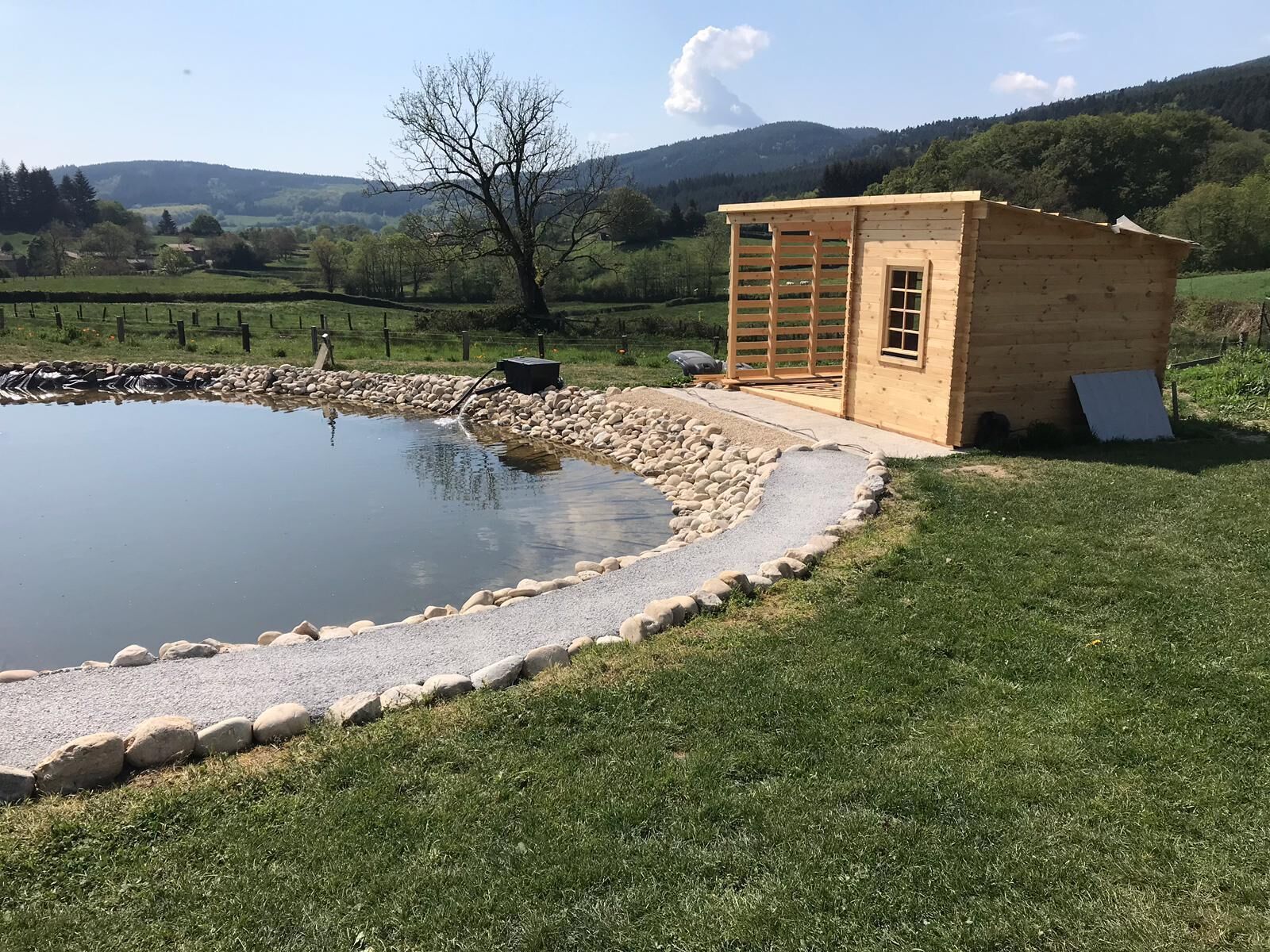 A wooden cabin with a small pond, surrounded by a stone border and grassy area, set in a rural landscape with hills in the background.