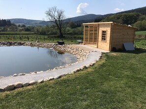 Une cabane en bois avec un petit étang, bordée d’un muret de pierres et entourée d’une zone herbeuse, située dans un paysage rural avec des collines en arrière-plan.