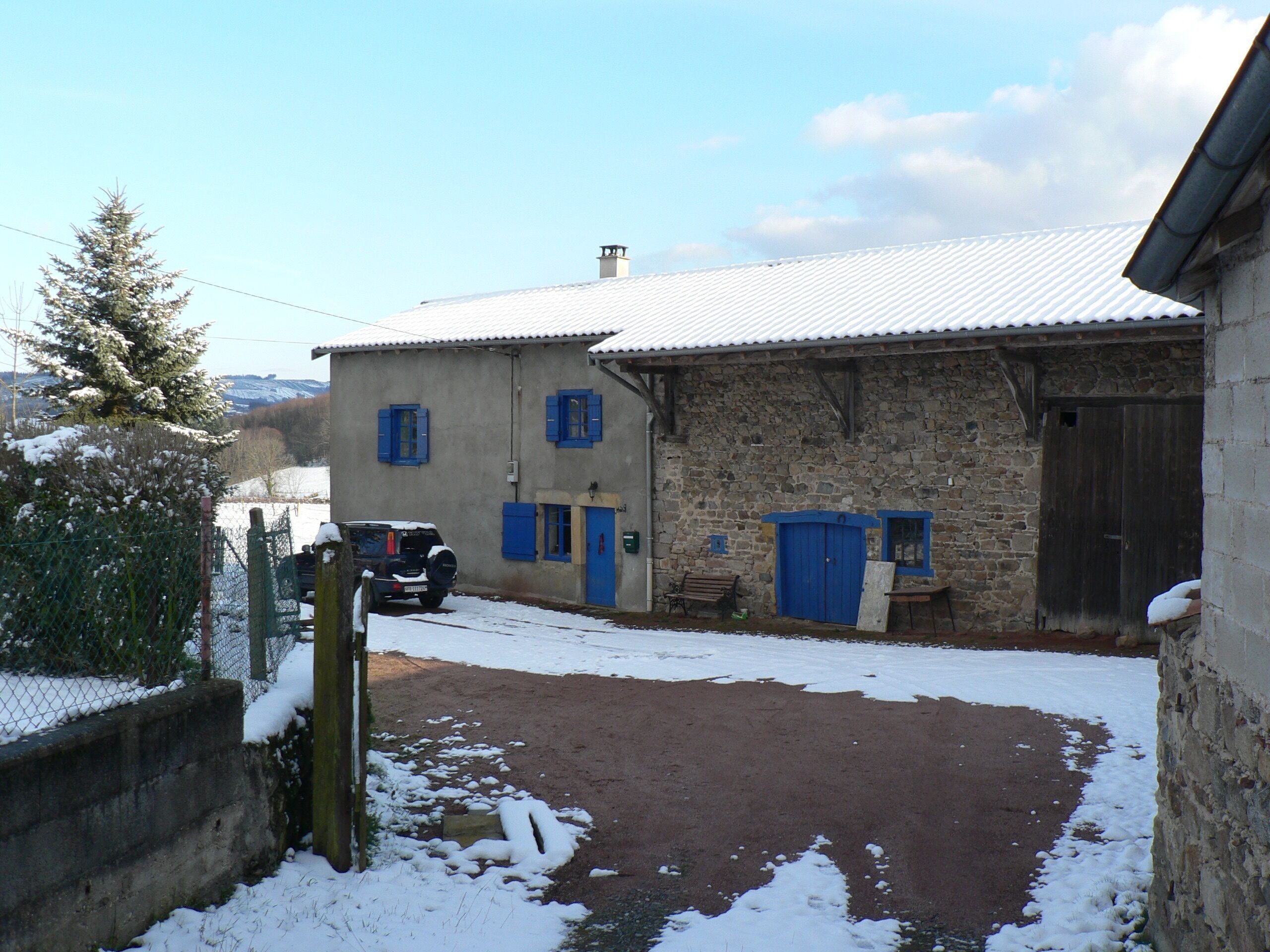 Un bâtiment en pierre avec des volets bleus, une voiture garée et un sol recouvert de neige.
