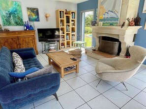 Sala de estar con sofá, sillón, mesa de centro, estantería, televisor, chimenea y vistas al exterior.