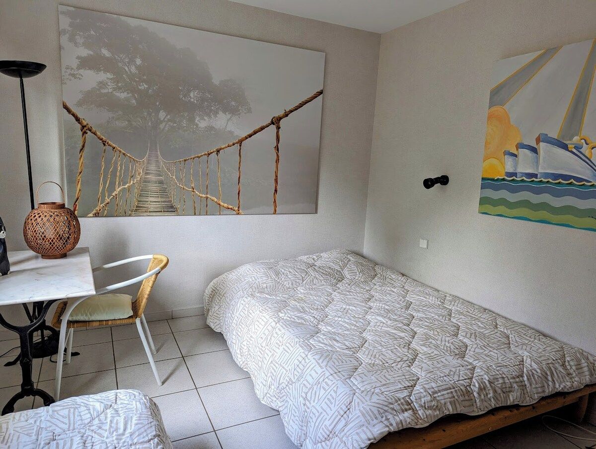 A bedroom with a bed, a chair, a table, a lamp, and artwork on the walls.