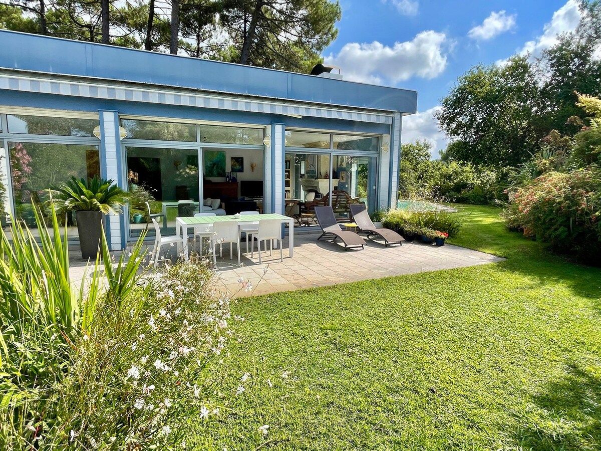 A modern house with a glass facade, a patio with outdoor furniture, and a well-maintained lawn.