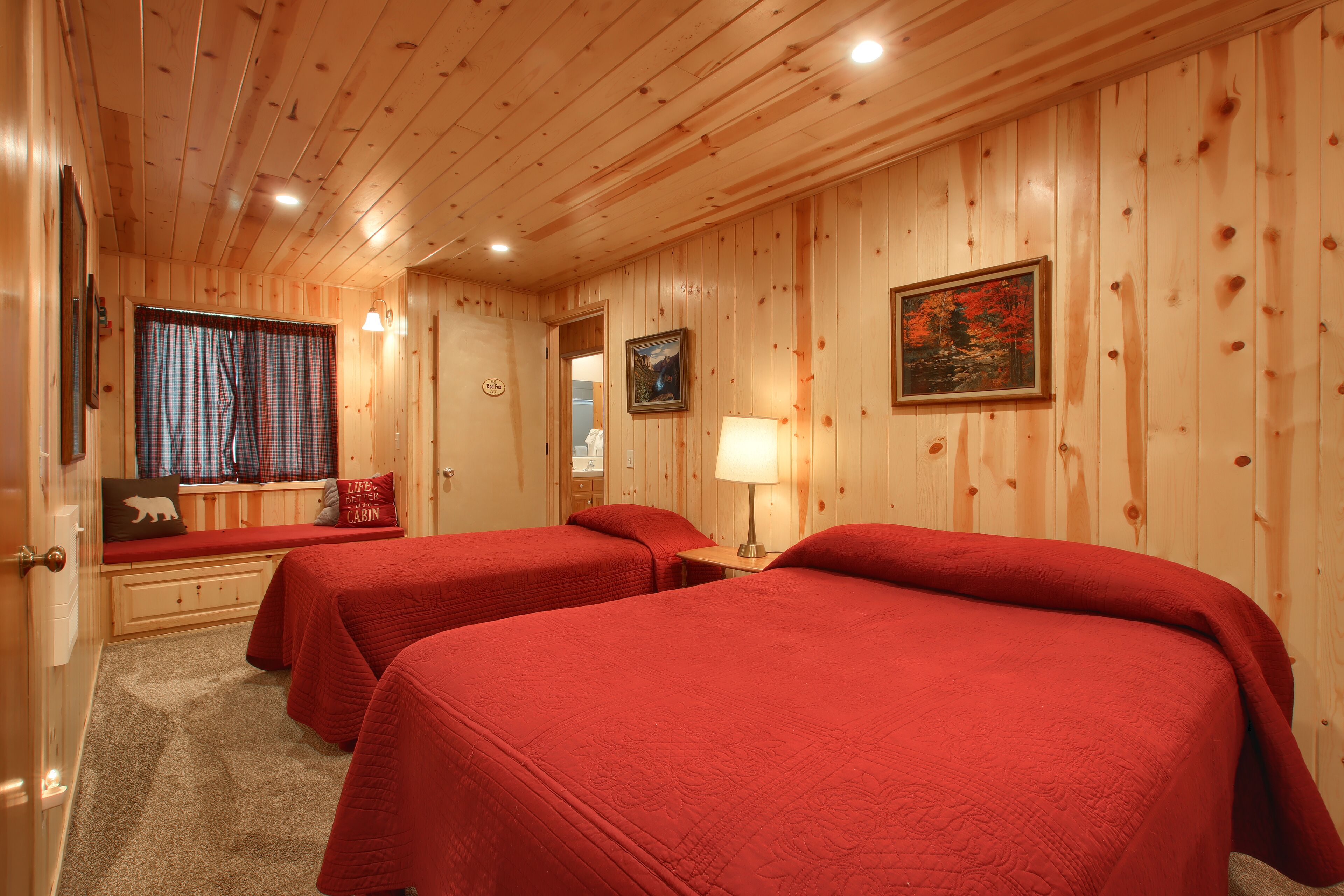 Ein blockhaushaltsähnliches Zimmer mit zwei Betten, Holzwänden, einer Bank mit Kissen, einem Gemälde und einer Lampe.