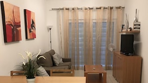 A living room with a sofa, a dining table with chairs, a TV on a wooden cabinet, and a potted plant. - Outstading Executive Apartment 150 metres from the beach (Albufeira)