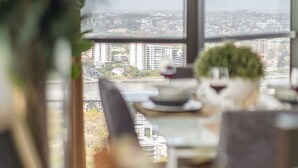A dining area with a table set for two, a potted plant, and a view of a cityscape through large windows. - Sky Homes on Queen (Brisbane)