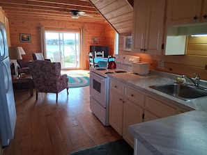 A cozy cabin interior with a wooden ceiling, a dining area, a kitchen with a stove and toaster, and a view through the glass door.