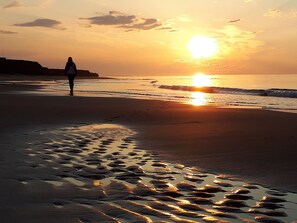A beach scene with footprints leading to the water, a person walking, and the sun setting over the ocean. - Sandcastles Beach House - Duneside: On PEI's Best Beach! (Darnley)