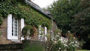 Una casa de piedra con paredes cubiertas de hiedra, contraventanas blancas y un jardín con diversas plantas y flores.