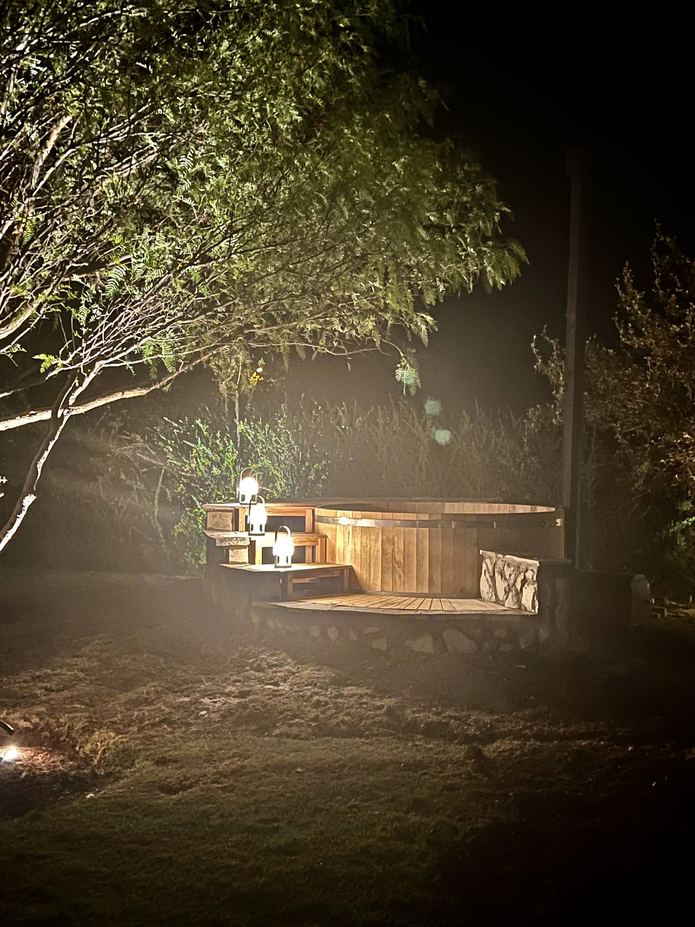 Un área de estar al aire libre con bancos y mesas de madera, rodeada de árboles y un muro de piedra.