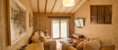 A cozy living room with a wooden ceiling, a large painting, a sofa set, a television, and a window with shutters.