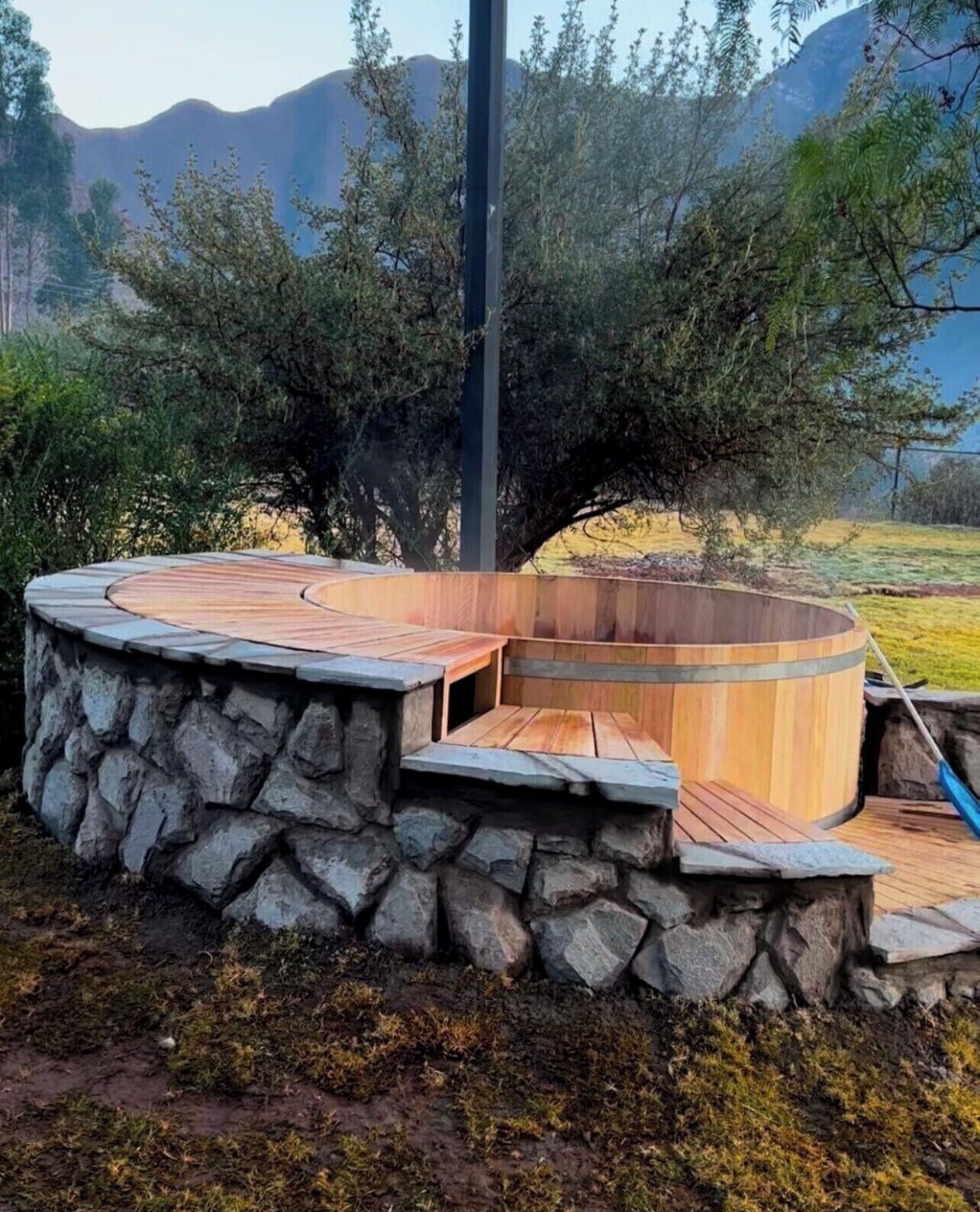 A circular wooden structure with stone steps, surrounded by a natural landscape with trees and mountains in the background.