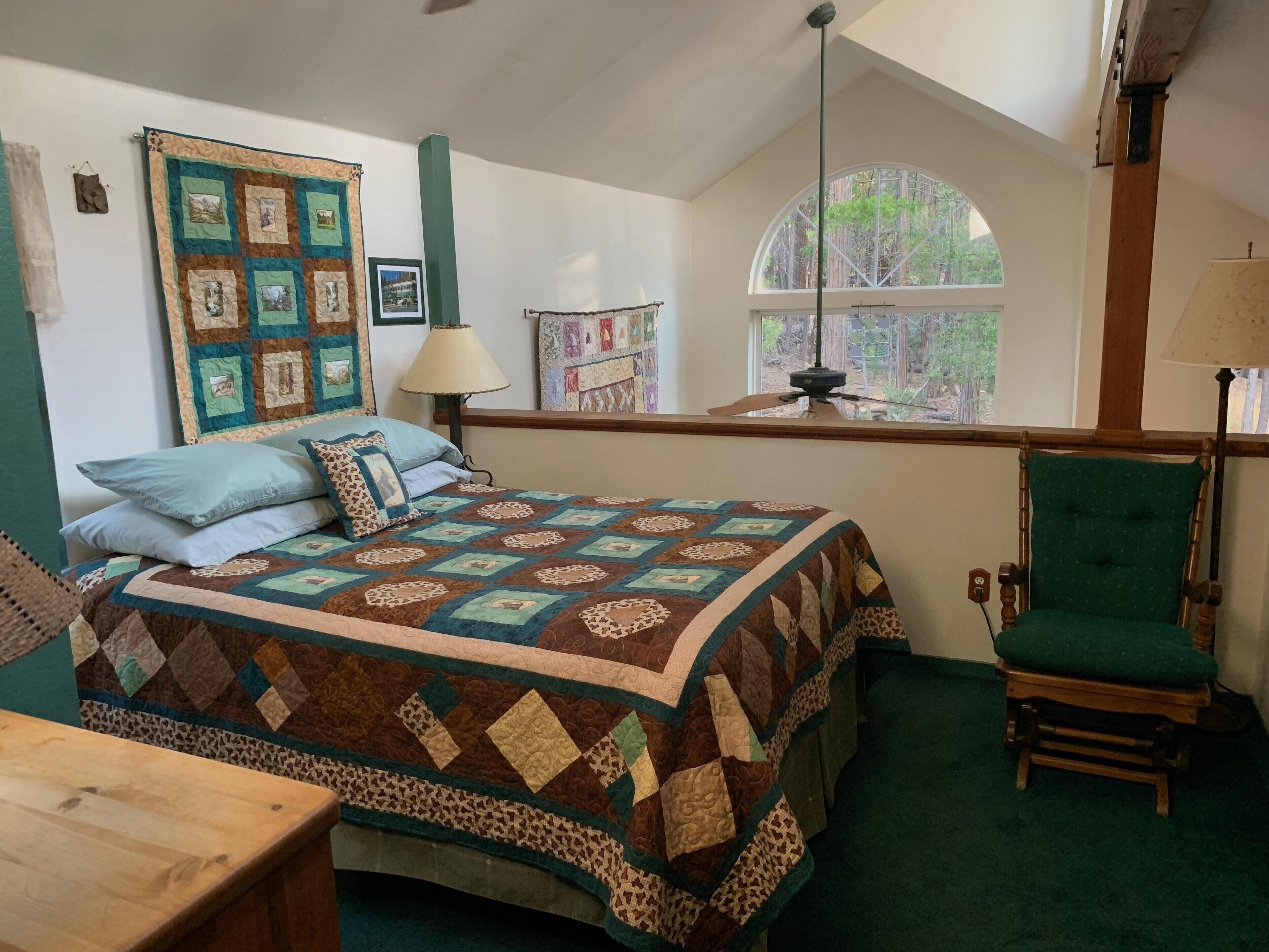 A bedroom with a large bed, a green armchair, a wooden table, and a window with a view of trees.