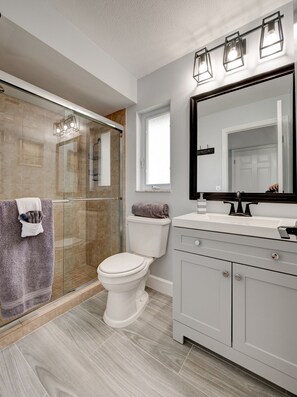 Een moderne badkamer met een glazen douchecabine, een wit toilet, een wastafelmeubel met spiegel en een raam boven de wastafel.