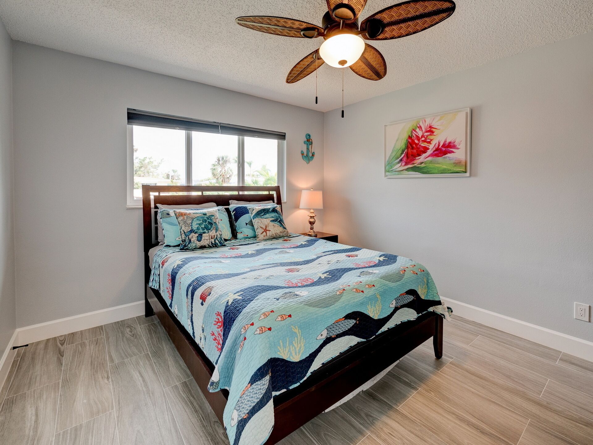 A bedroom with a bed, a ceiling fan, a window, a lamp, and a painting on the wall.