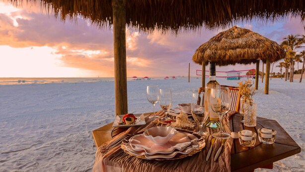 A beachside dining setup with a table set for two, featuring plates, glasses, and a candle.