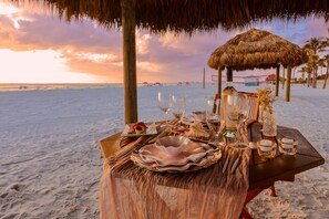 Een eetgedeelte aan het strand, met een tafel gedekt voor twee personen, inclusief borden, glazen en een kaars.