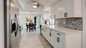 A modern kitchen with white cabinetry, a stainless steel refrigerator, a dining area with a table and chairs, and a gray-tiled backsplash.