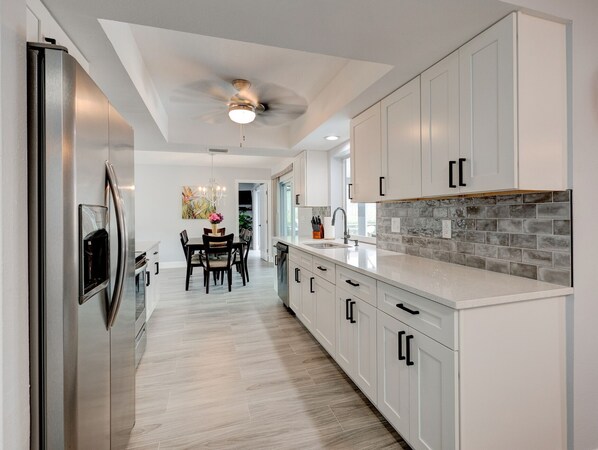 A modern kitchen with white cabinetry, a stainless steel refrigerator, a dining area with a table and chairs, and a gray-tiled backsplash.