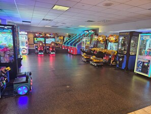 Un salón de juegos interior con diversas máquinas recreativas, incluyendo juegos de carreras y de garra con disco, y un paseo accesible para sillas de ruedas.