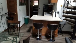 A compact kitchen with a central island, two bar stools, a glass dining table, and a trash bin.