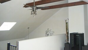 A room with a sloped ceiling, exposed wooden beams, a ceiling fan, a wall-mounted TV, and a partial view of a staircase.