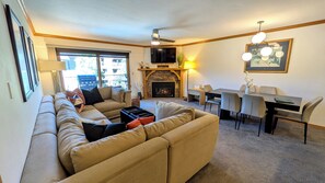 Ein Wohnzimmer mit einer Couch, einem Kamin, einem Esstisch mit Stühlen, einem Deckenventilator und einem an der Wand befestigten Fernseher.