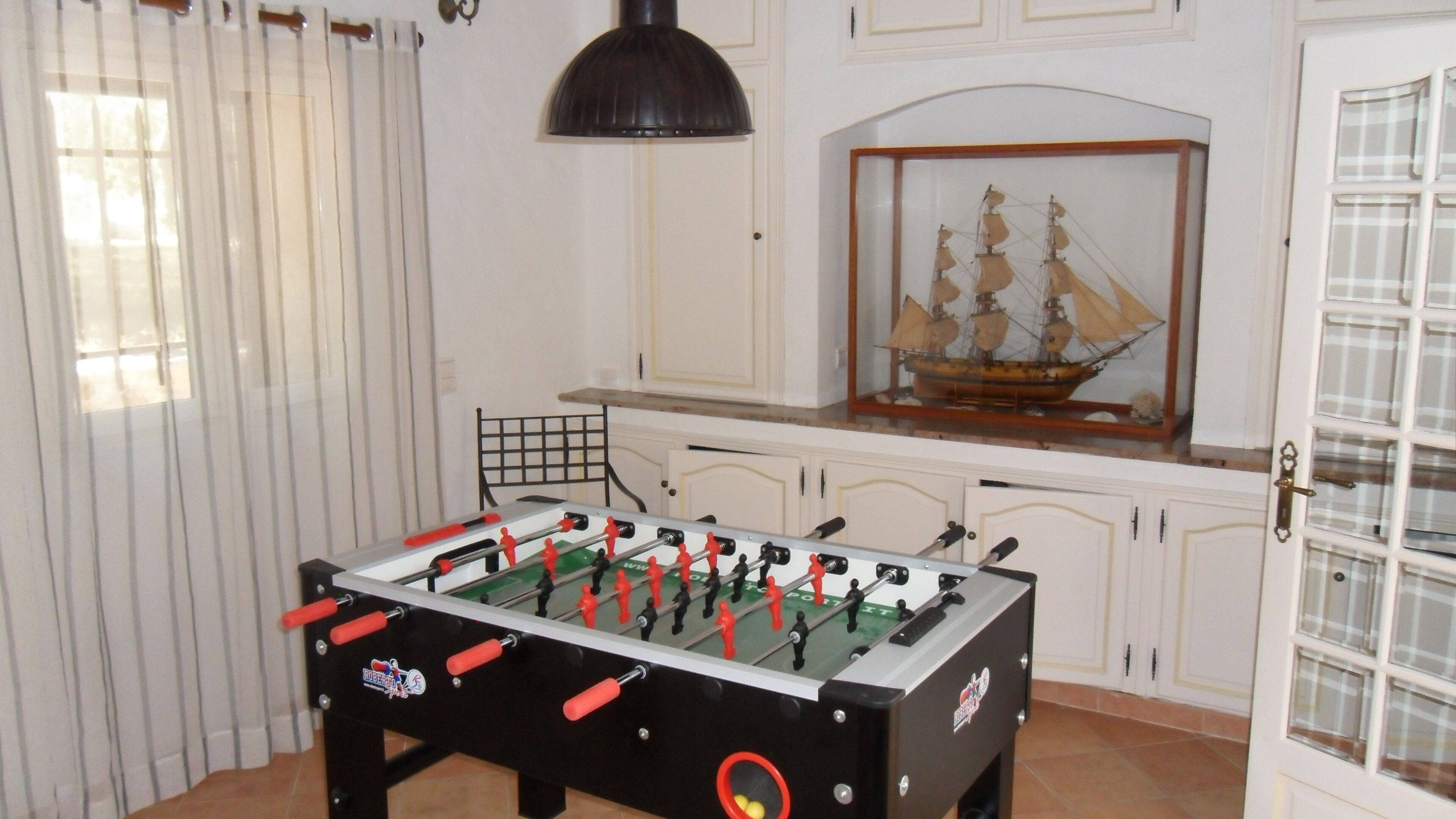 A foosball table in a room with white cabinets, a ship model, and a window with curtains.