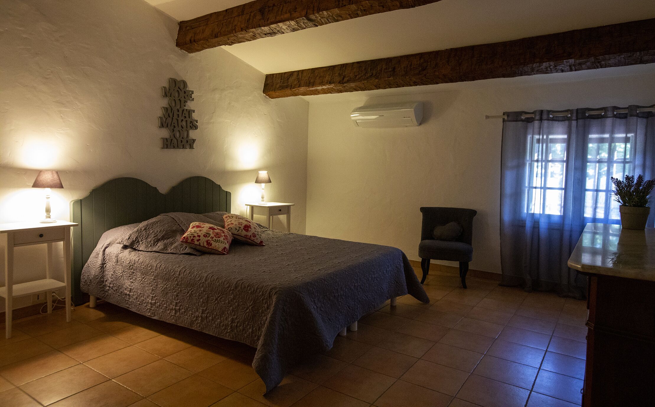 Une chambre à coucher comprenant un lit, deux tables de chevet, une chaise, un bureau, une plante en pot et une fenêtre avec des rideaux.