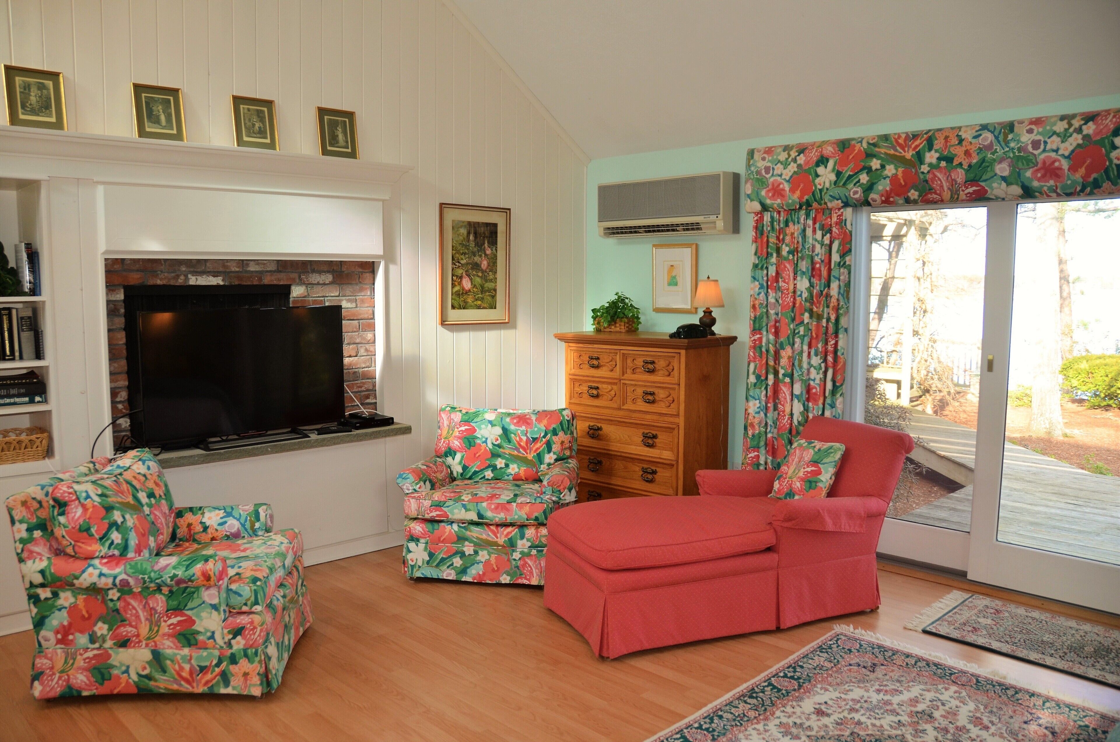 A living room with a fireplace, a television, floral-patterned chairs, a red sofa, and a wooden dresser.
