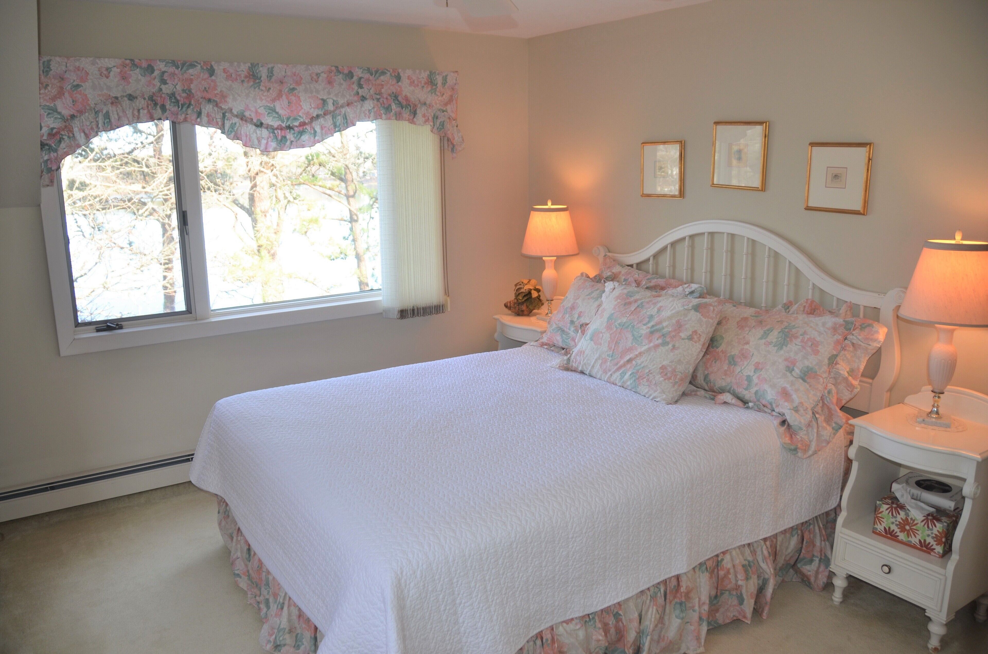 Una camera da letto con un letto, due comodini con lampade, una finestra con tenda a fiori e tre quadri appesi al muro.