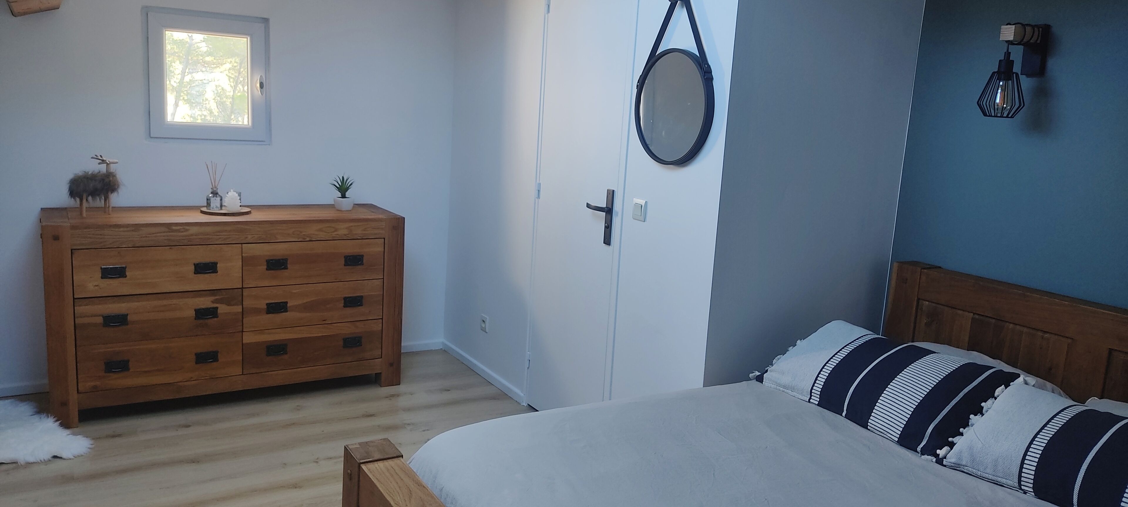 Un dormitorio con un aparador de madera, una cama con almohadones a rayas, un espejo redondo y una ventana pequeña.