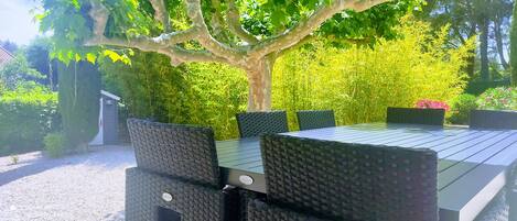A rectangular outdoor table with four chairs, surrounded by lush greenery and a tree.