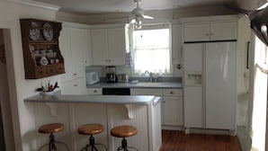 A kitchen with white cabinets, a central island with stools, a microwave, a coffee maker, and a refrigerator. - 2 Bedroom Lovingly Restored 1925  Cottage 1 1/2 Blocks From Beach (Tybee Island)